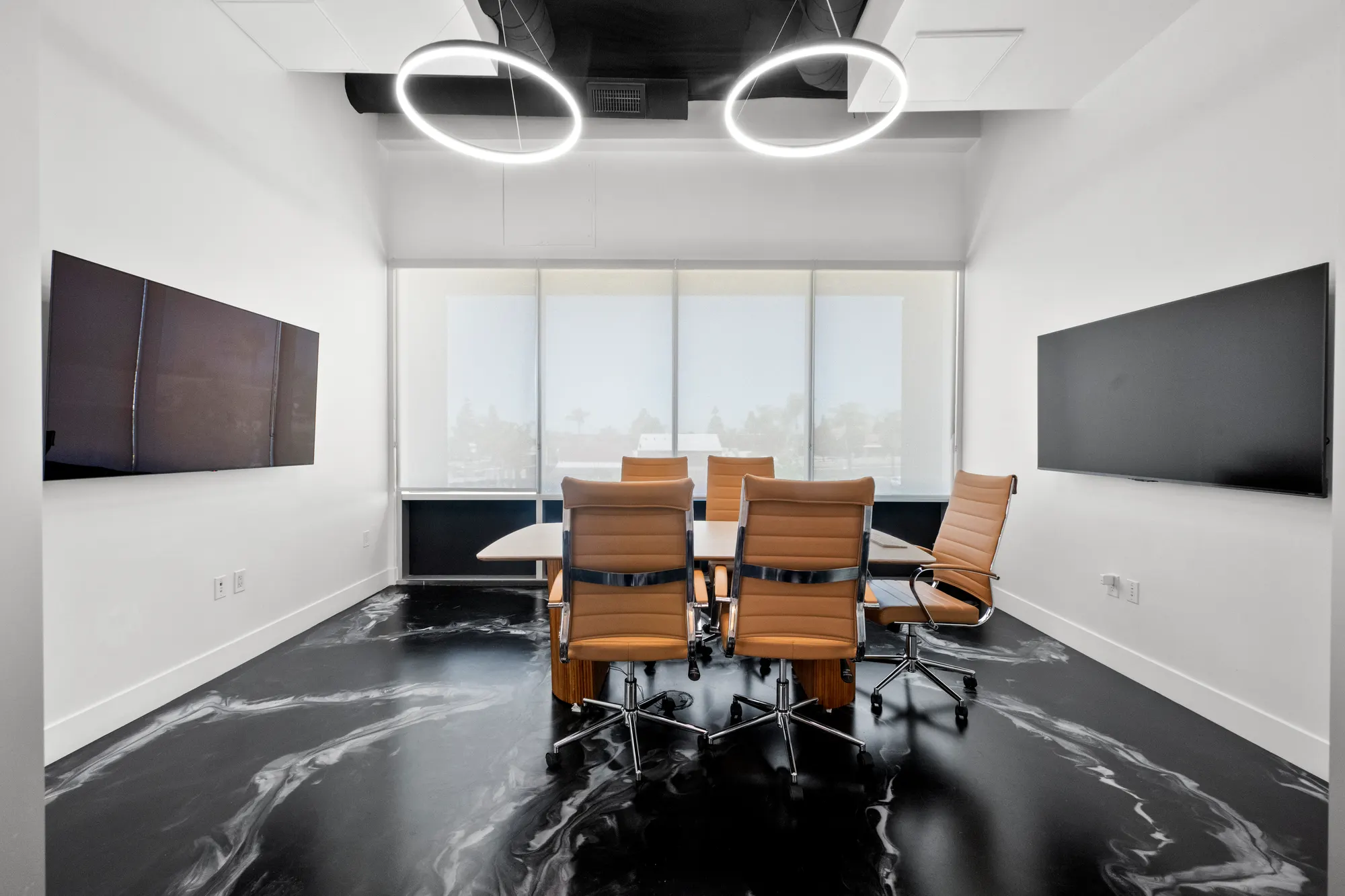 Sala de juntas con paredes de vidrio, una mesa larga, sillas de piel color camel, dos lámparas circulares en el techo, pantallas en la pared y piso de mármol negro.