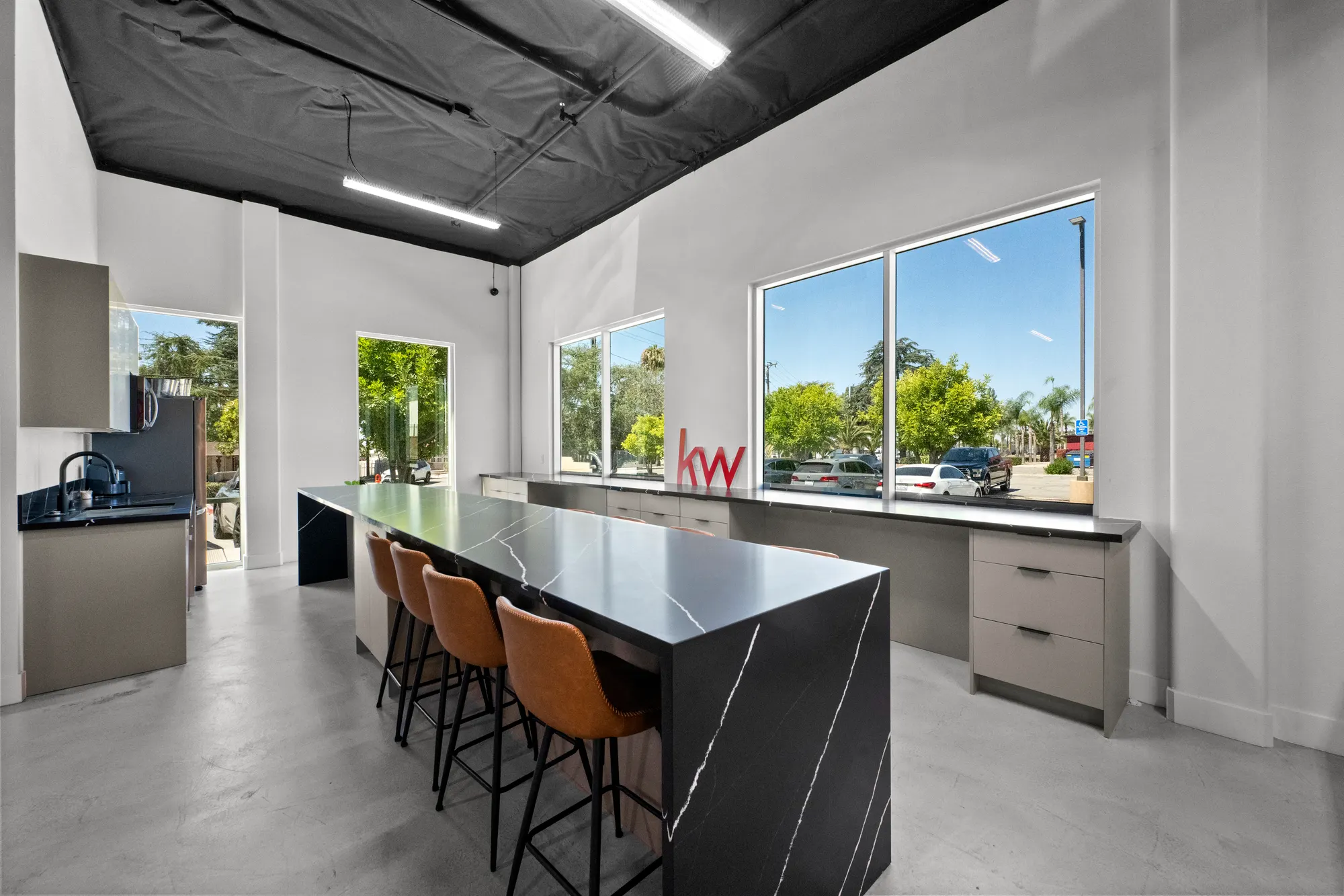 Cocina y sala de descanso con una isla de mármol negro, bancos de piel y una fila de ventanas con letras rojas de kw en el alféizar y árboles afuera.