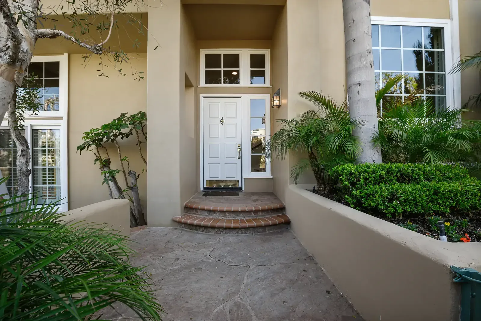 Front entry of a beige stucco home with a white door, curved brick steps and palms on either side